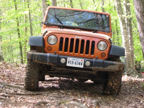 1979 Ford Bronco - An orange Jeep Wrangler is shown off-road, navigating a wooded trail with muddy tires.