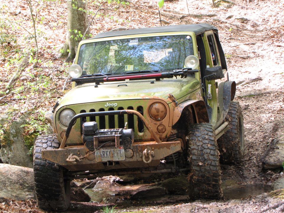 Green Jeep Wrangler with winch and off-road tires on a muddy trail. Virginia license plate RKGRVL