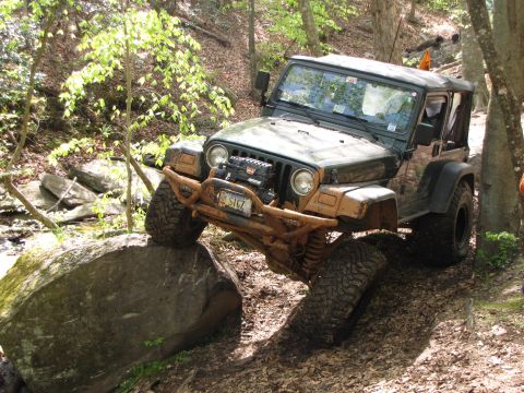 1979 Ford Bronco - A Jeep Wrangler navigates a challenging off-road obstacle on a dirt path surrounded by trees.