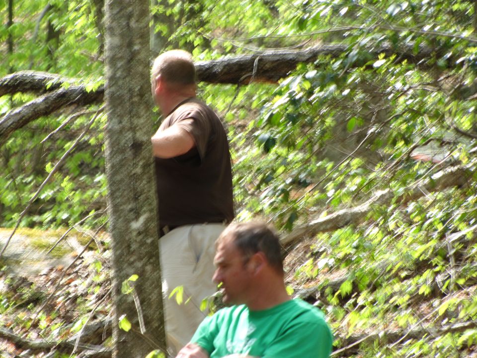 Two men are in a wooded area. One man in a brown shirt and tan pants stands behind a tree. Another man in a green shirt is in the foreground