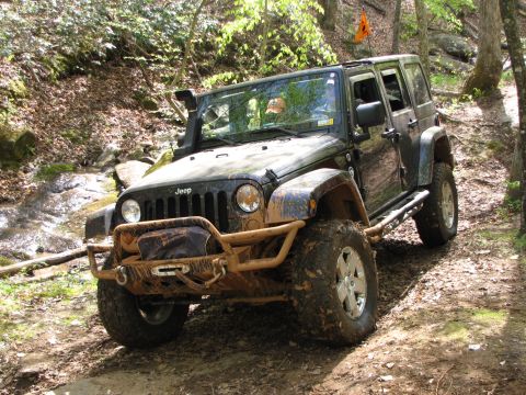 1979 Ford Bronco - A muddy Jeep Wrangler navigates a wooded trail with an orange flag visible on its antenna.