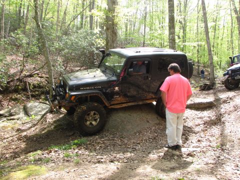 1979 Ford Bronco - A black Jeep Wrangler Rubicon is shown off-roading on a dirt trail in a wooded area, with another vehicle visible in the background.