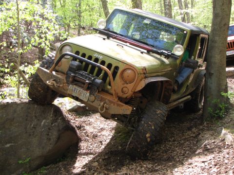 1979 Ford Bronco - A Jeep navigates a challenging off-road obstacle on a dirt path surrounded by trees.