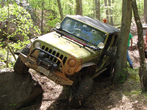 1979 Ford Bronco - A green Jeep Wrangler navigates a challenging off-road course, with one tire on a large rock and the vehicle angled against a tree.