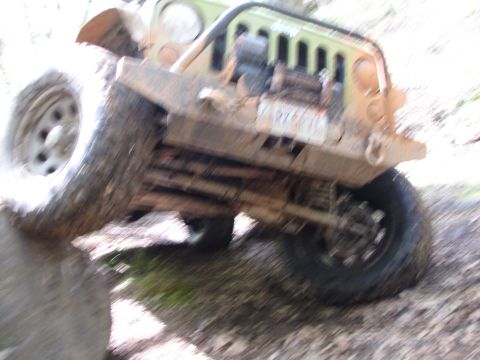 1979 Ford Bronco - A muddy Jeep Wrangler navigates an off-road obstacle, with its front tire climbing over a large rock.