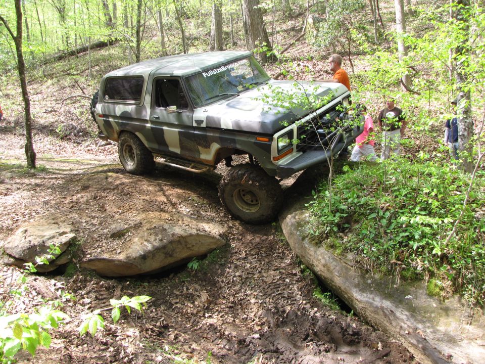 Camouflaged 1978 Ford Bronco navigating a rocky, wooded trail. FullsizeBronco.com sticker on windshield
