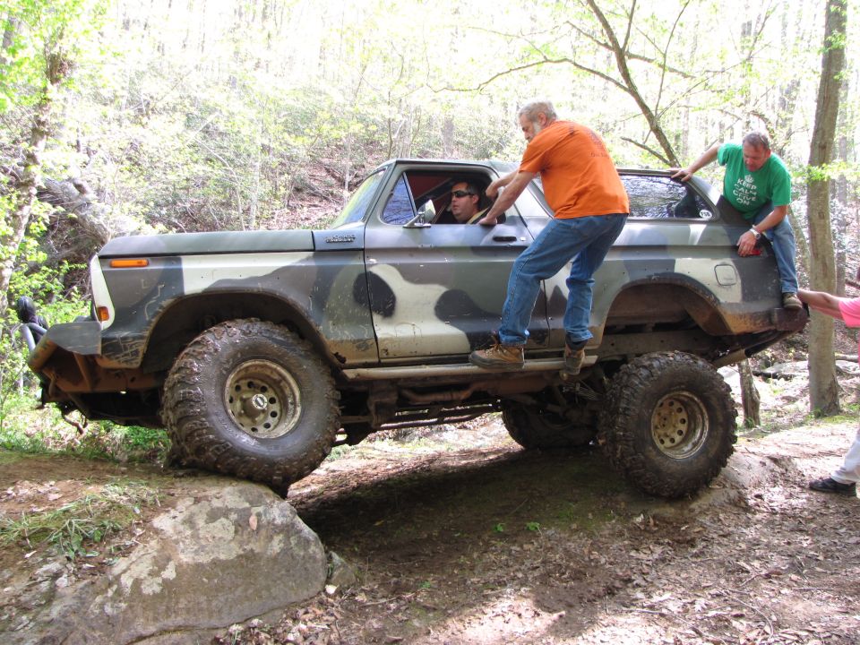 Camouflaged Ford Bronco with lifted suspension and off-road tires navigating rocks, with people climbing on it
