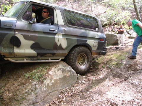 1979 Ford Bronco - A driver navigates a camouflage Ford Bronco over large rocks on a wooded trail.