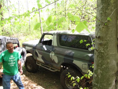 1979 Ford Bronco - A 1979 Ford Bronco with camouflage paint is parked in a wooded area, with a man standing beside it.