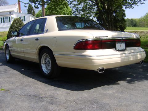 1996 Mercury Grand Marquis - The rear of a cream-colored sedan is parked on an asphalt surface, with a house and trees in the background.