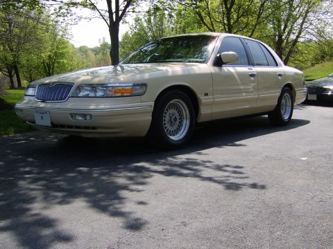 1996 Mercury Grand Marquis - A tan sedan with a chrome grille and wire-mesh wheels is parked outdoors on a sunny day.