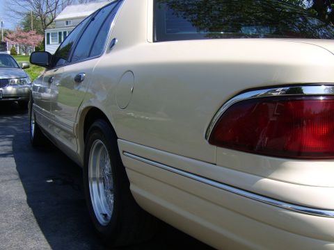 1996 Mercury Grand Marquis - A cream-colored sedan is parked on the street next to a house, with another car in the background.