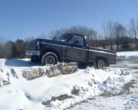 1985 Ford F-150 - Driver-side view of a black regular-cab Ford pickup with a chrome grille perched atop a snow-covered mound on a sunny winter day. Snow bank…