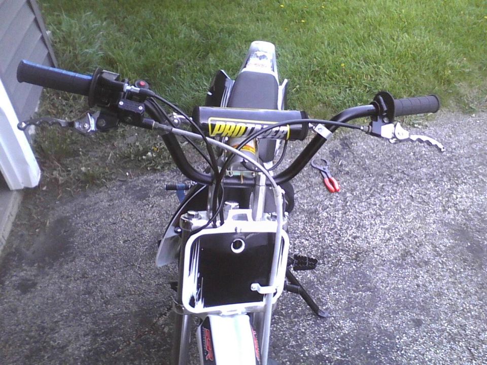 Dirt bike handlebars with "PRO TAPER" branding, black grips, and brake lever. Front number plate is visible