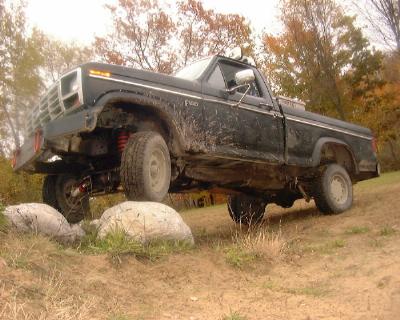 1985 Ford F-150 - A mud-splattered black Ford F-Series regular-cab long-bed pickup flexes over a row of boulders, front driver's wheel lifted clear of the gr…