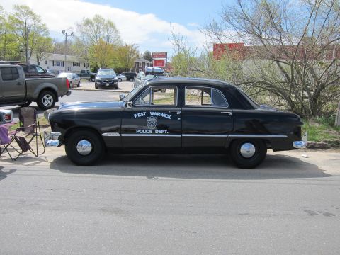 1991 Ford LTD Crown Victoria - Side view of a black West Warwick Police Dept. sedan with a roof spotlight parked on pavement beside folding chairs.