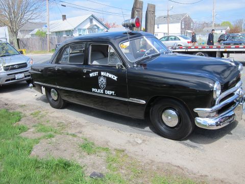 1991 Ford LTD Crown Victoria - black West Warwick Police Dept. sedan with a single roof siren parked on a gravel shoulder near houses and a car trailer.