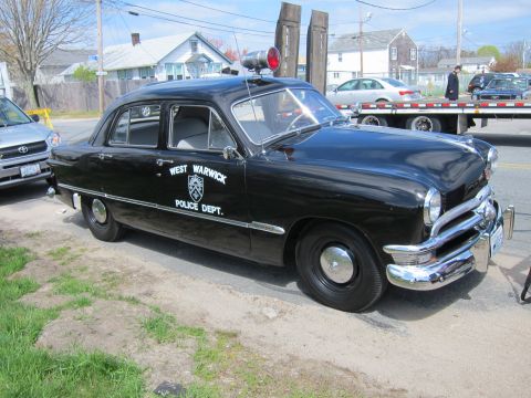 1991 Ford LTD Crown Victoria - Profile of a black West Warwick Police Dept. sedan with a roof siren parked on a dirt edge near a flatbed trailer.