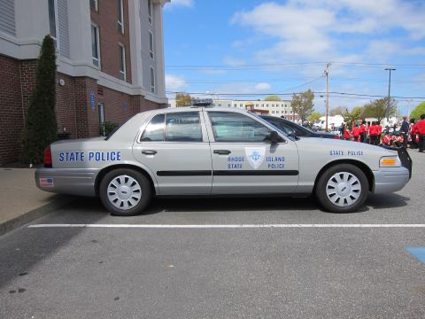 1991 Ford LTD Crown Victoria - silver Rhode Island State Police Ford Crown Victoria sedan shown in side profile with a marching crowd in red behind it.