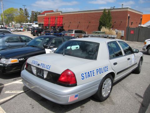 1991 Ford LTD Crown Victoria - silver State Police Ford Crown Victoria shown from the rear quarter with light bar and flag decal, parked by a brick building.