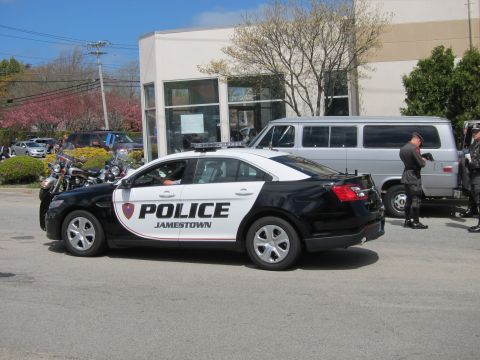 1991 Ford LTD Crown Victoria - black-and-white Jamestown Police Ford Taurus Interceptor sedan parked near a building with a van and motorcycle behind it.