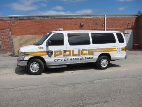 1991 Ford LTD Crown Victoria - Side view of a white Ford E-Series police van marked POLICE CITY OF HACKENSACK with gold striping against a brick wall.