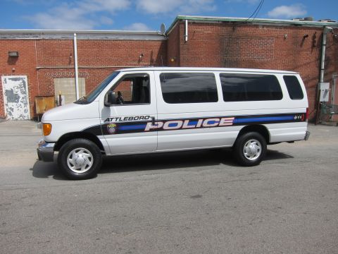 1991 Ford LTD Crown Victoria - Side profile of a white Ford E-Series police van marked ATTLEBORO POLICE with red, white and blue striping by a brick building.