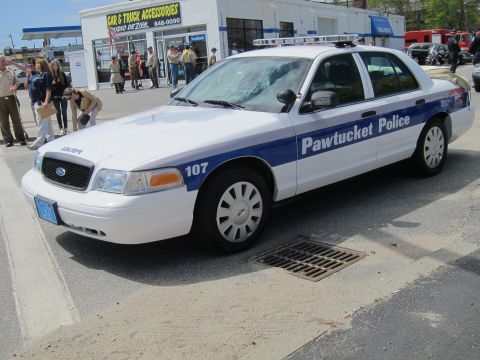 1991 Ford LTD Crown Victoria - white and blue Ford Crown Victoria marked Pawtucket Police, unit 107, parked front-three-quarter with people on the sidewalk behind.