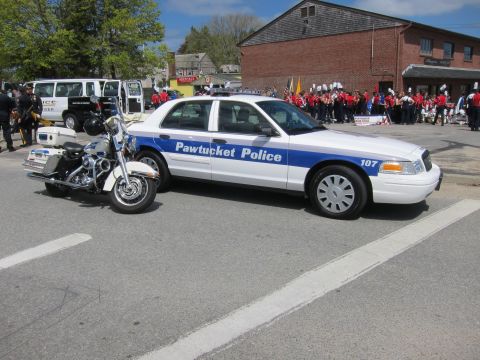 1991 Ford LTD Crown Victoria - white and blue Pawtucket Police Ford Crown Victoria, unit 107, parked beside a police motorcycle near a marching band.