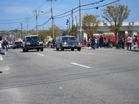 1991 Ford LTD Crown Victoria - black vintage sedan and a dark SUV roll down a wide street as people watch from the curb on a clear day.