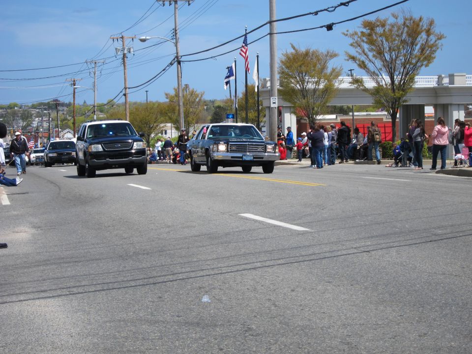 Black vintage sedan and black SUV driving down a street at a parade