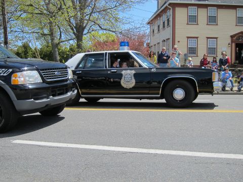 1991 Ford LTD Crown Victoria - black vintage Ford police sedan unit 45 with a blue beacon drives past spectators and a tan house at a parade.