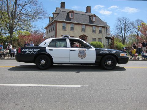 1991 Ford LTD Crown Victoria - black-and-white Springfield Police Ford Crown Victoria unit 7 marked Emergency 911 drives past a tan colonial house.