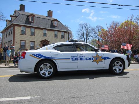 1991 Ford LTD Crown Victoria - white Norfolk County Sheriff Dodge Charger with gold star badge drives past spectators holding US flags at a parade.