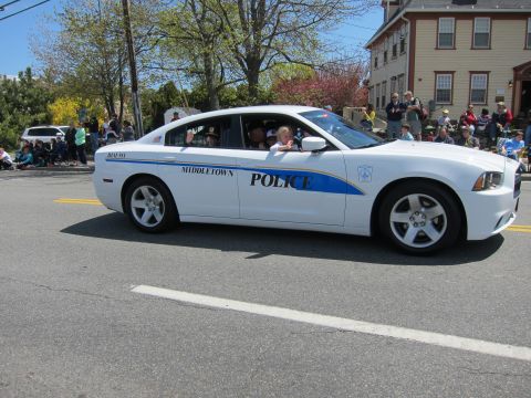 1991 Ford LTD Crown Victoria - white Dodge Charger Middletown Police sedan with blue striping drives a parade route past a crowd and a brick building.