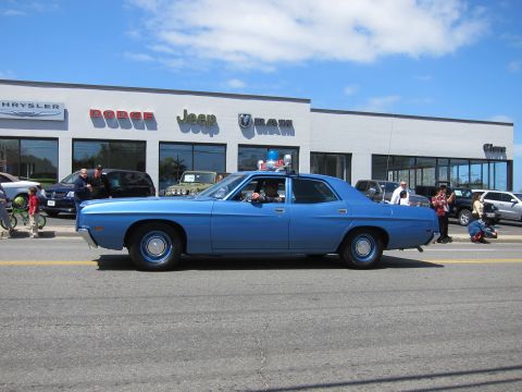 1991 Ford LTD Crown Victoria - blue vintage four-door sedan with a single roof light drives past a Dodge Jeep Ram dealership.