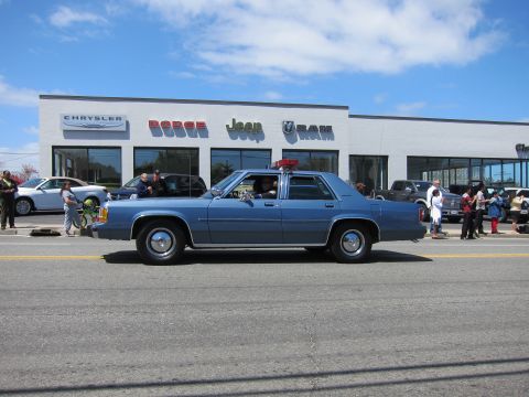1991 Ford LTD Crown Victoria - blue Ford LTD Crown Victoria sedan with a roof light bar drives past a Chrysler Dodge Jeep Ram dealership.