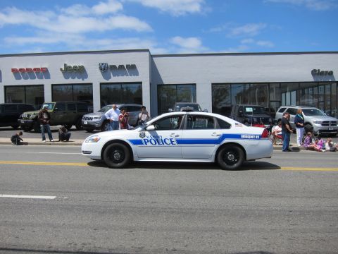 1991 Ford LTD Crown Victoria - white Chevrolet Impala marked Police with a blue stripe drives past a Dodge Jeep Ram dealership as crowds watch.
