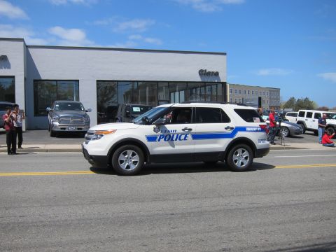 1991 Ford LTD Crown Victoria - white Ford Explorer marked Police with a blue stripe drives past a dealership as spectators line the street.