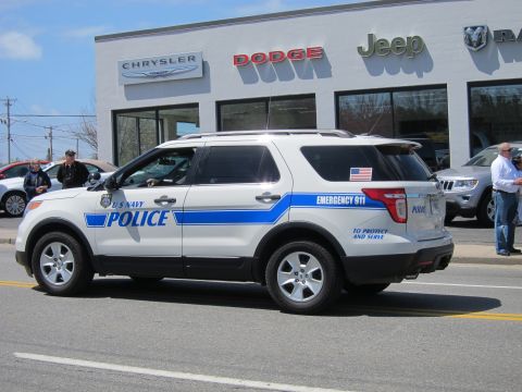 1991 Ford LTD Crown Victoria - white Ford Explorer police SUV lettered "U S Navy Police" and "Emergency 911" drives past a Chrysler-Dodge-Jeep dealership.