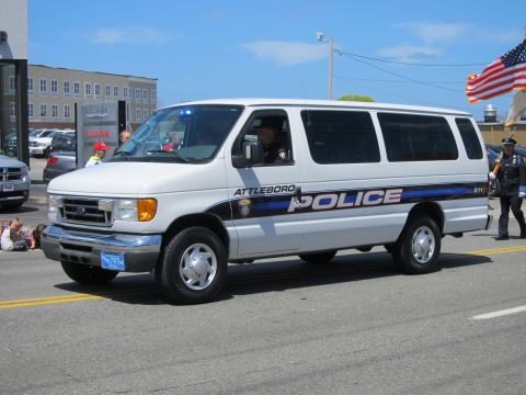 1991 Ford LTD Crown Victoria - white Ford E-Series Attleboro Police van rolls down a street with onlookers and flags on a sunny day.