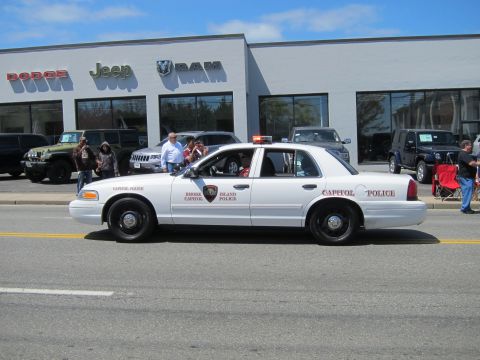 1991 Ford LTD Crown Victoria - white Ford Crown Victoria Capitol Police cruiser passes a Dodge-Jeep-RAM dealership as people watch from chairs.