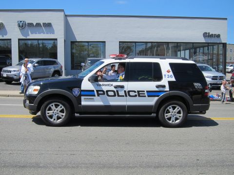 1991 Ford LTD Crown Victoria - black-and-white Warwick Police Ford Explorer SUV passes a RAM dealership with onlookers seated curbside.