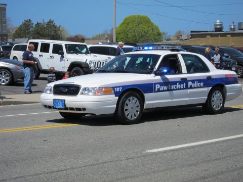 1991 Ford LTD Crown Victoria - white Pawtucket Police Ford Crown Victoria cruiser, number 107, drives past parked vehicles with lights active.