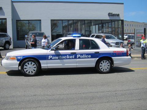1991 Ford LTD Crown Victoria - white-and-blue Pawtucket Police Ford Crown Victoria, number 107, passes a dealership with spectators on the sidewalk.