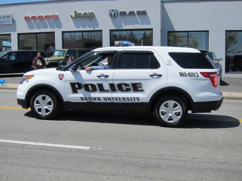 1991 Ford LTD Crown Victoria - white Ford Explorer SUV lettered "POLICE Brown University" with roof lightbar drives past a Jeep and Ram car dealership.