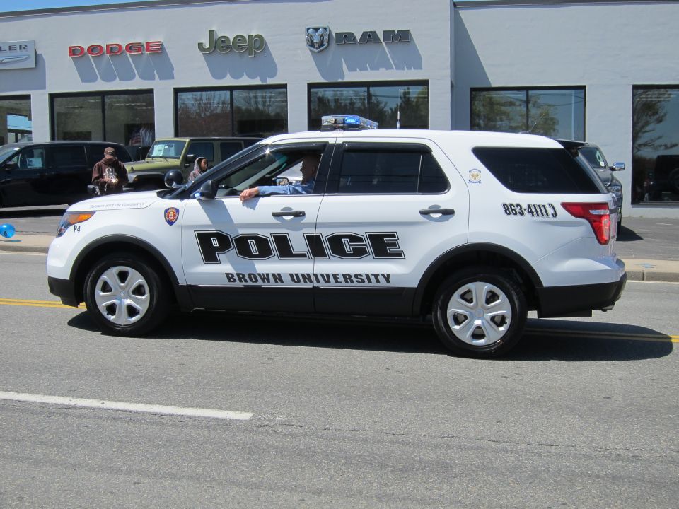 White Ford Explorer SUV marked Brown University Police on a road by a Dodge/Jeep/Ram dealership