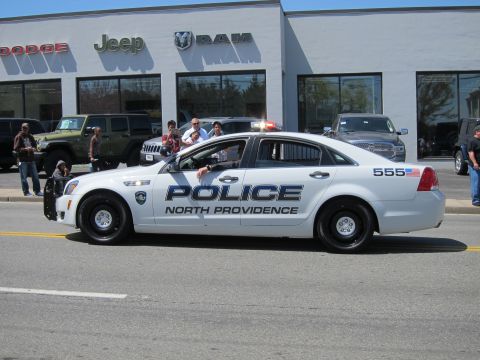 1991 Ford LTD Crown Victoria - white North Providence Police Chevrolet Caprice cruiser numbered 555 drives past a Jeep and Ram dealership with spectators.