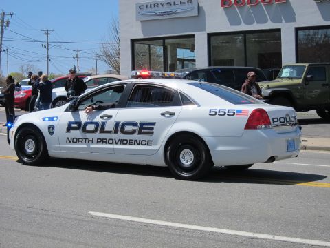 1991 Ford LTD Crown Victoria - white North Providence Police Chevrolet Caprice numbered 555 with lit roof lights drives past a Chrysler/Dodge dealership.