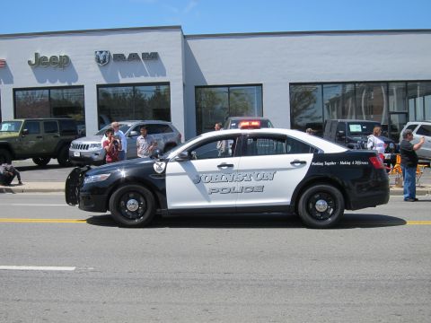 1991 Ford LTD Crown Victoria - black and white Ford Taurus cruiser lettered "Johnston Police" with roof lightbar passes a Jeep and Ram dealership with onlookers.